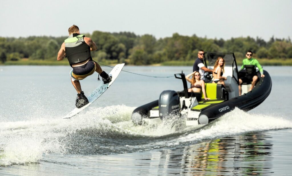 A group of friends on a boat looking back at a man jumping in the air on a wakeboard